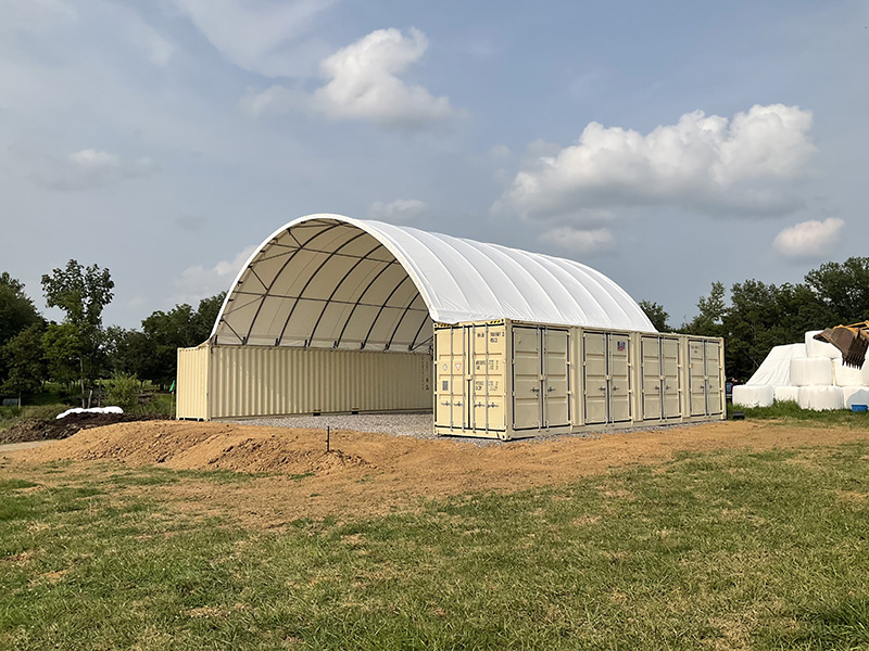 Two beige shipping containers support a large white shipping container canopy for covering and storing equipment and tools. 