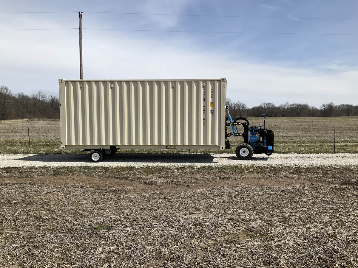 Blue Mule mover transporting a beige 20 foot shipping container along a rural gravel driveway.