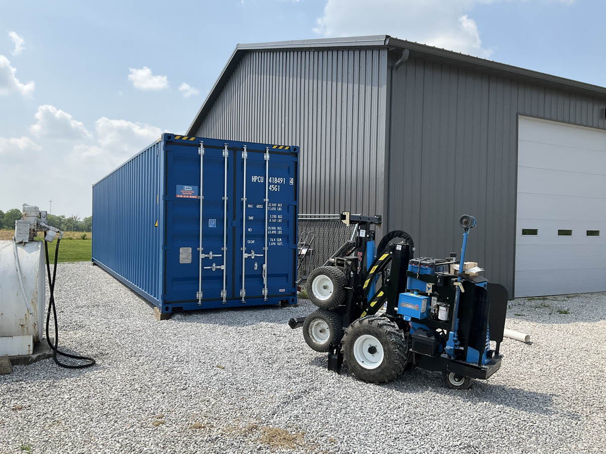 Mule forklift performing shipping container delivery beside a blue 40 foot shipping container set on a gravel pad next to a gray metal shop with a roll up door.