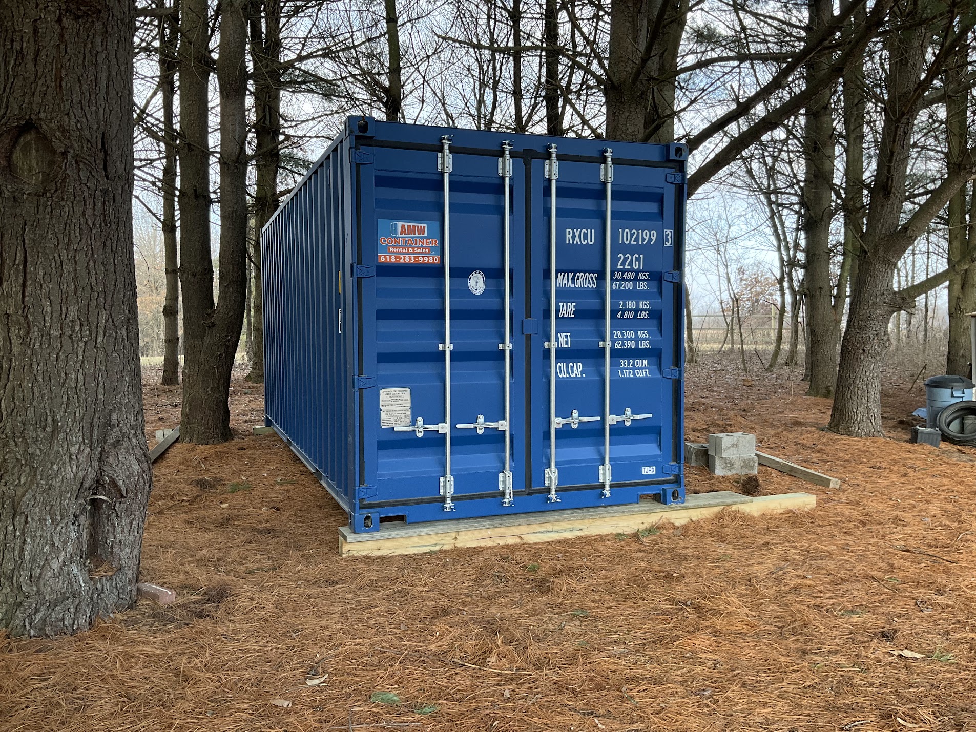 Blue 20-foot wind- and water-tight shipping container placed on a level foundation at a wooded property.
